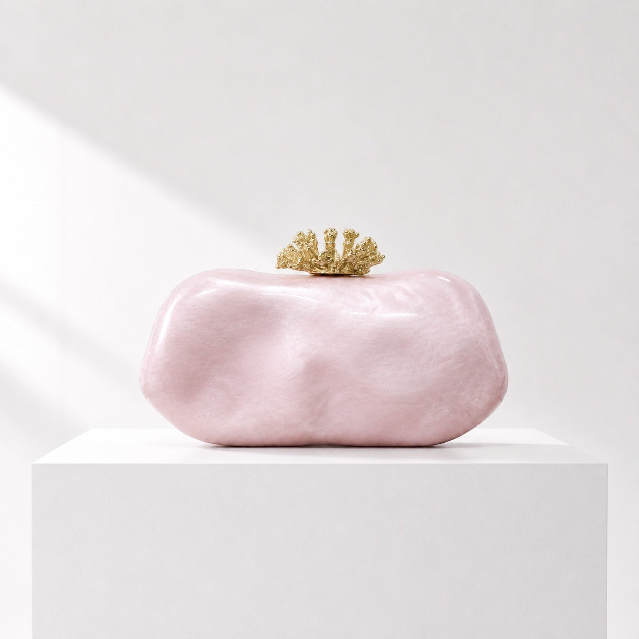 Lunéra de Paris pink acrylic oval clutch bag with a decorative gold-tone clasp on a white pedestal.