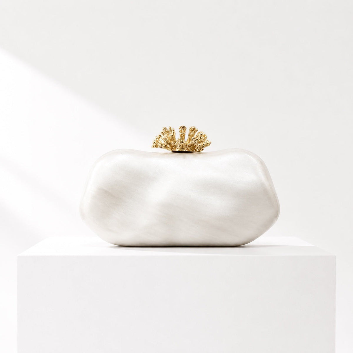 Lunéra de Paris oval evening clutch in shimmering white with a decorative gold floral clasp on a white pedestal